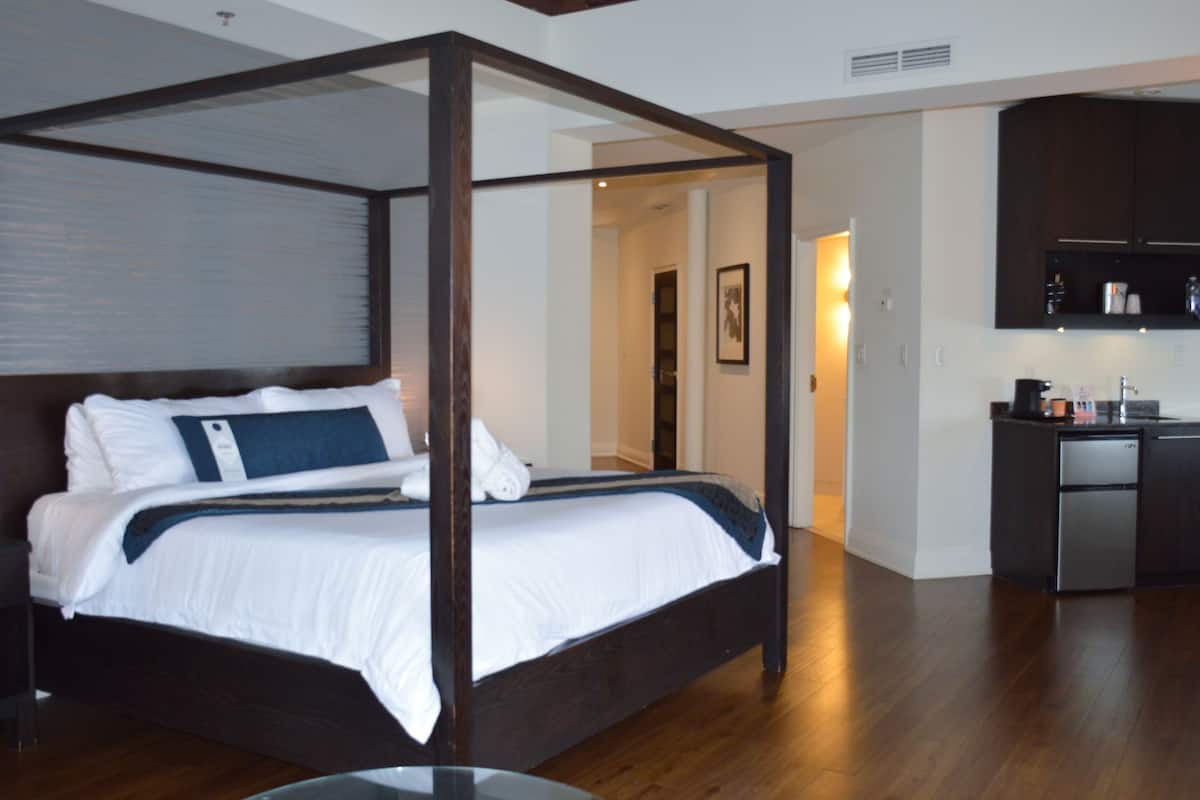 A four-poster bed with white bedding and a dark headboard, a kitchenette with dark cabinets, and a wooden floor.