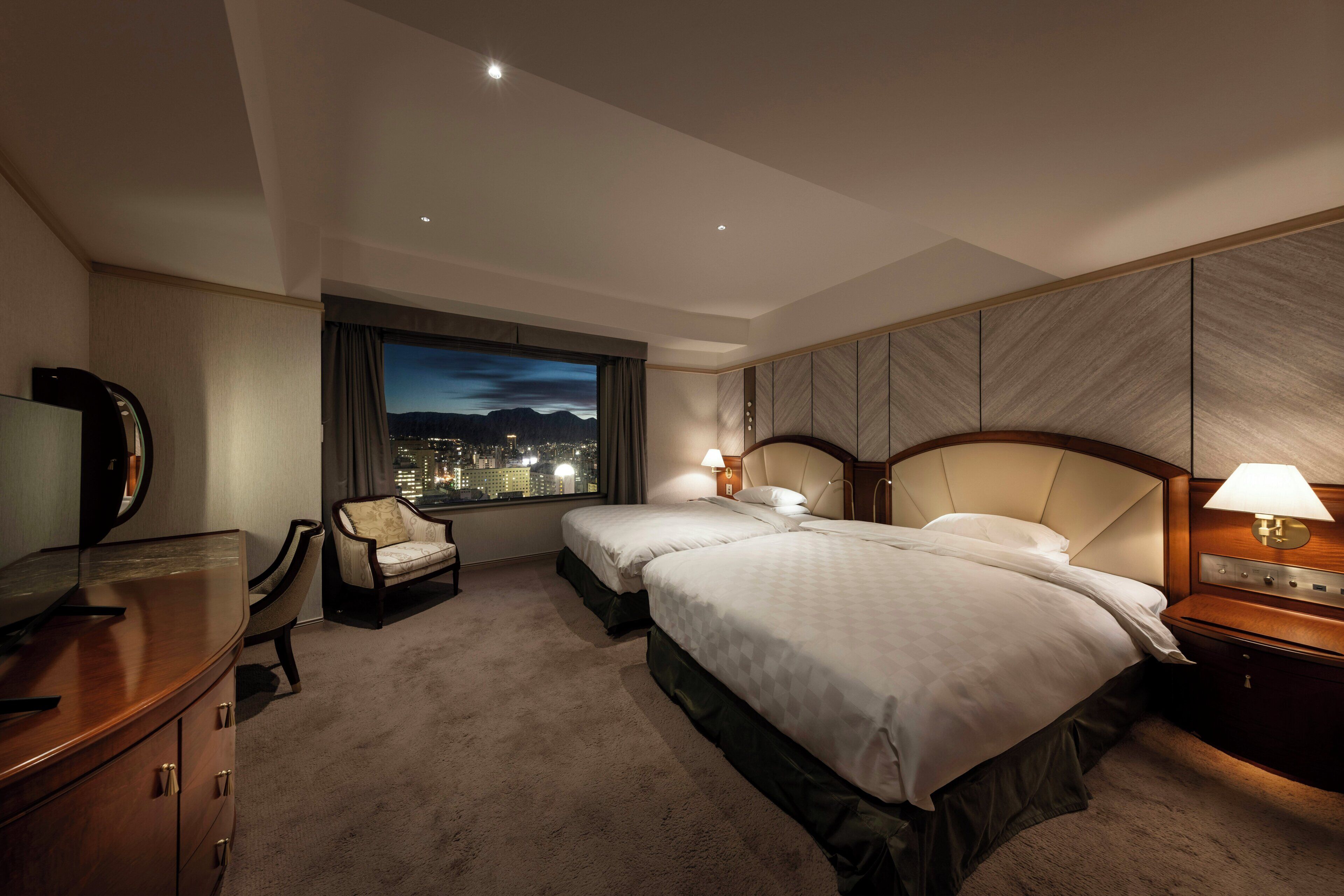 A hotel room with two beds, a desk, a chair, a TV, and a window with a city view.