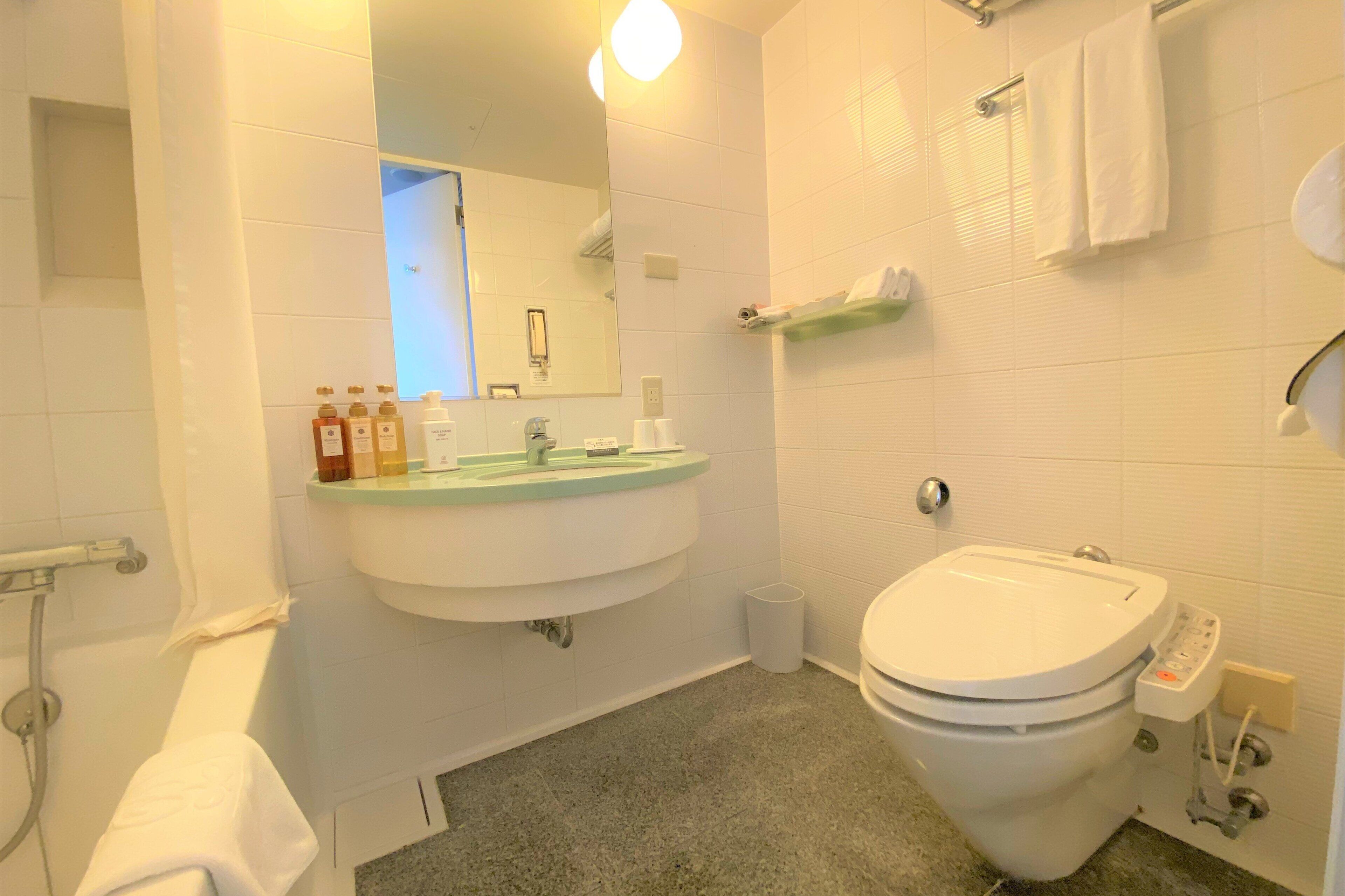 Un baño con lavamanos blanco, espejo e inodoro. Hay artículos de aseo en el lavamanos, un perchero y un bote de basura.