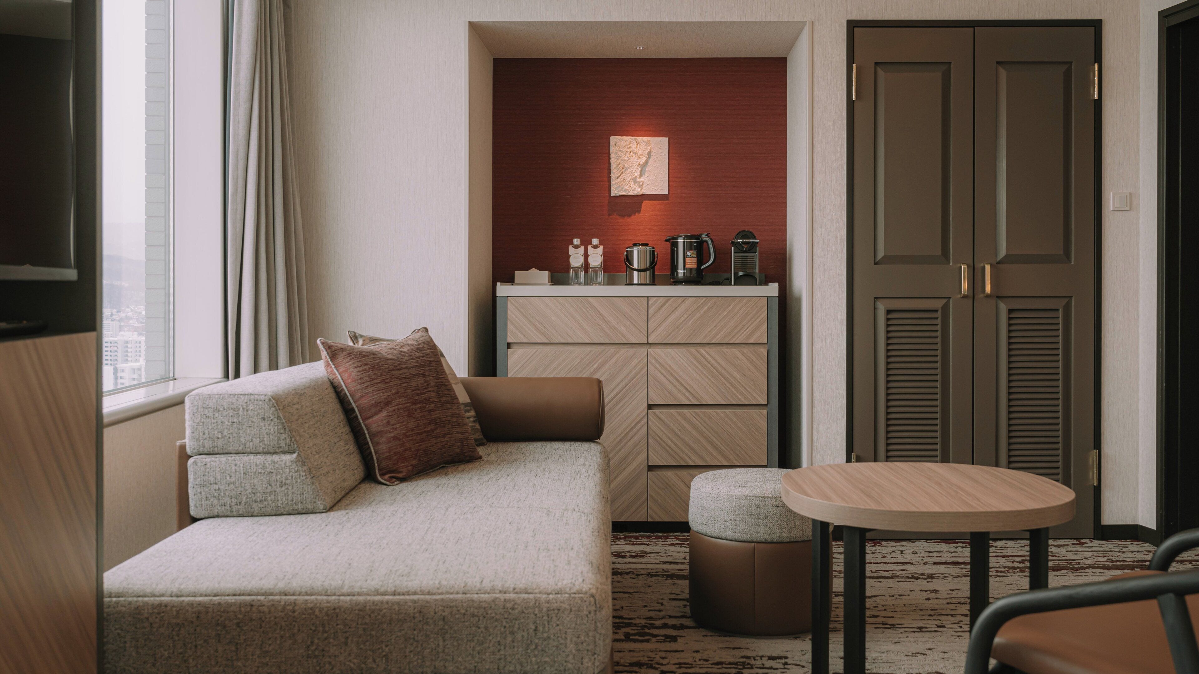A modern hotel room with a sofa, a small table, and a kitchenette area.