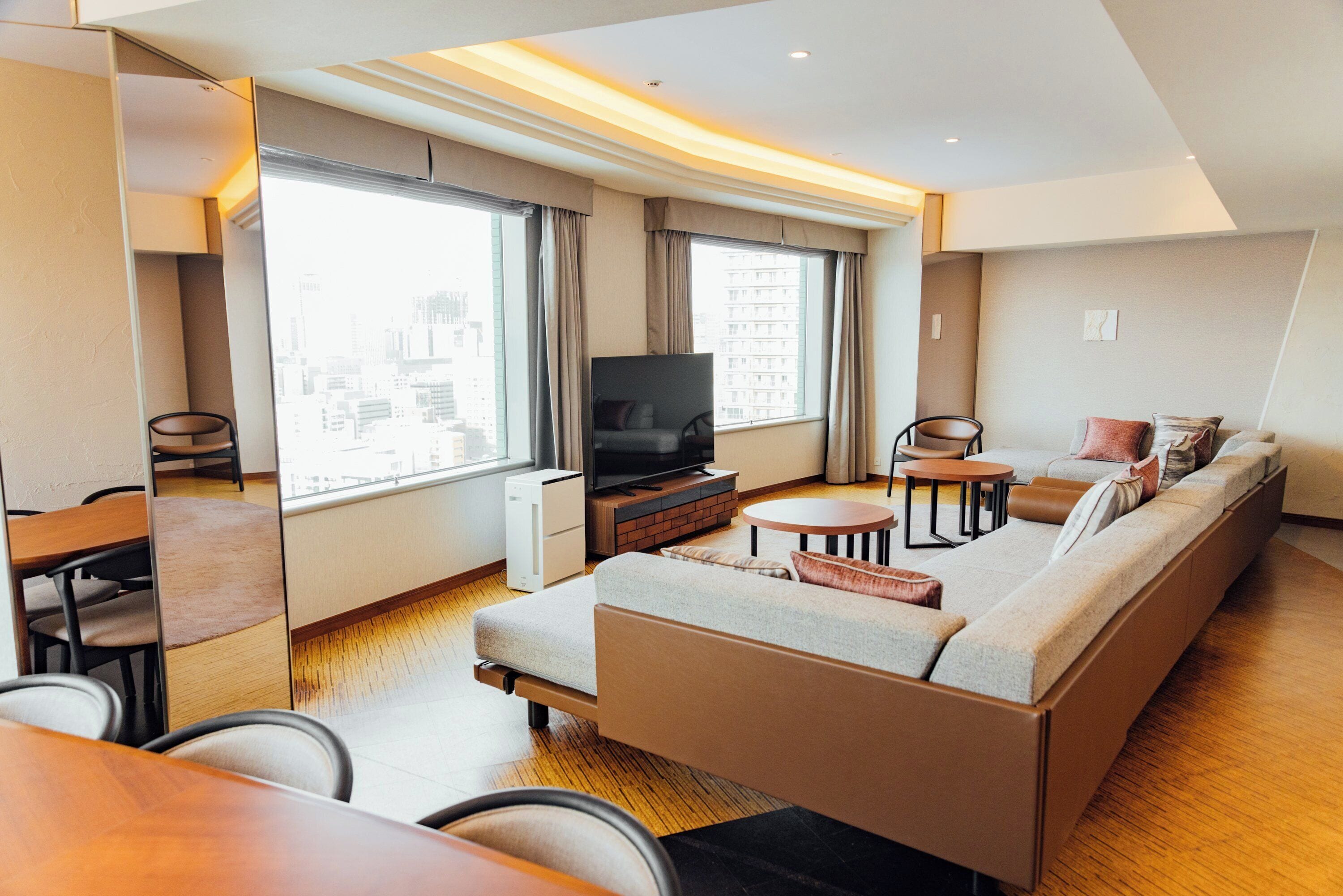 A modern hotel room with a sofa, TV, dining table, and large windows offering city views.