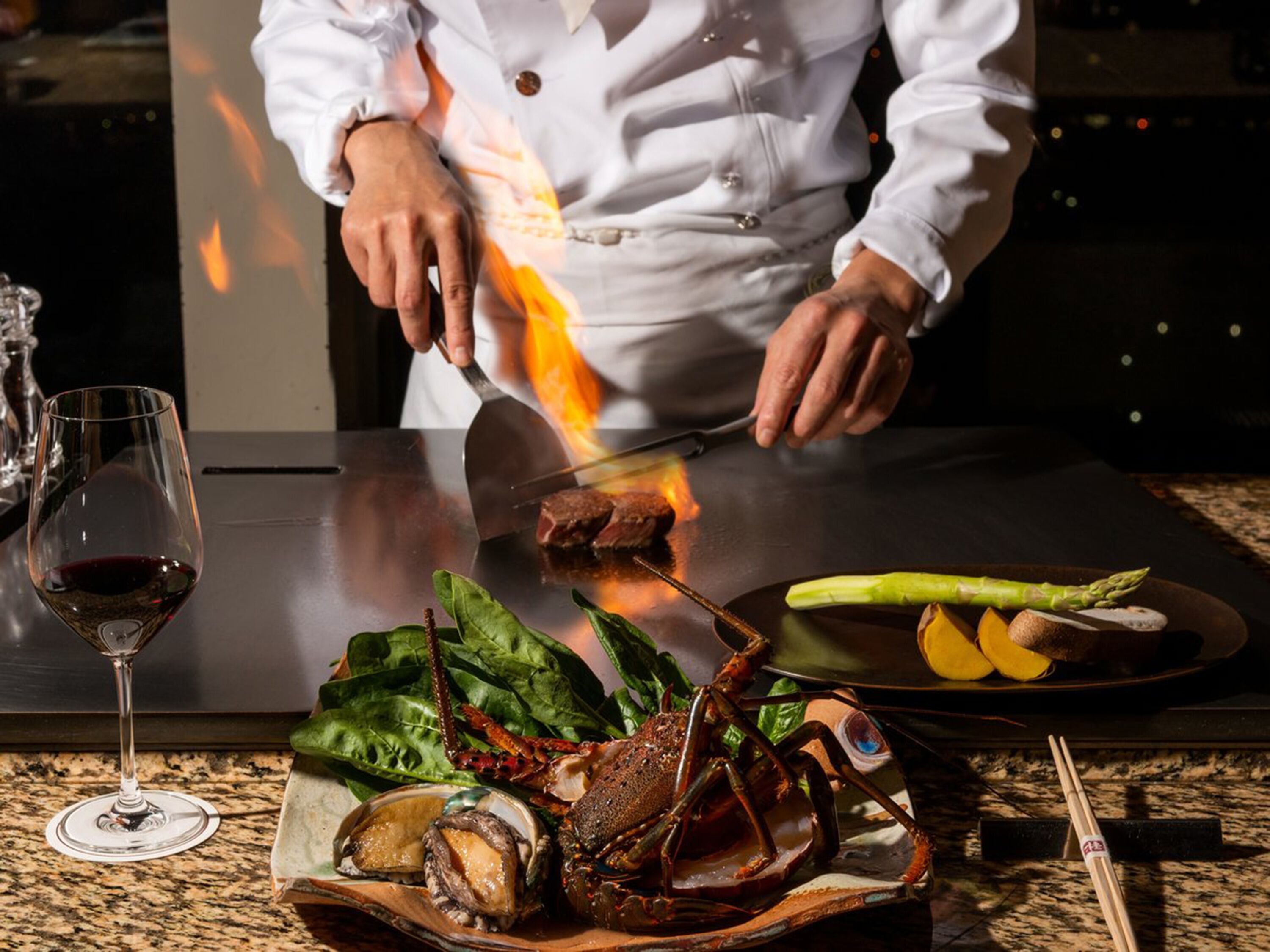 Un chef asando langosta en una mesa con un plato de verduras y una copa de vino.