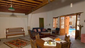 Ein gemütliches Wohnzimmer mit einem Holzesstisch, Stühlen, einer Couch und einer Bank. Ein Kamin, ein Fenster und eine Tür zu einem Außenbereich sind vorhanden.