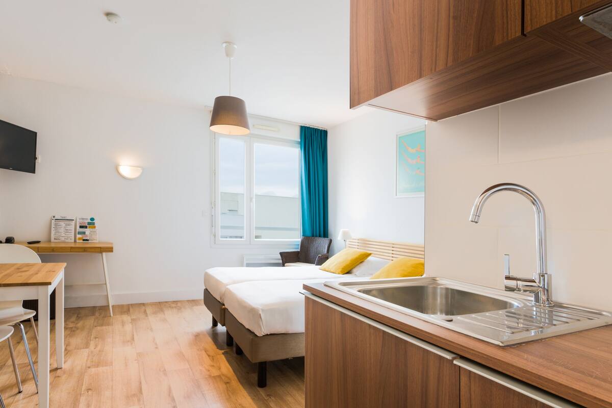 A compact hotel room with a kitchenette, a bed with white bedding, a desk with a chair, and a window with blue curtains.