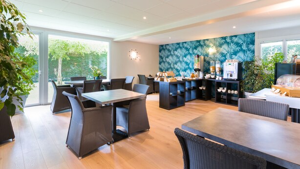 A modern dining area with wicker furniture, a kitchen counter, and a large window offering a view of greenery. - Appart'Hotel Victoria Garden Pau (Pau)