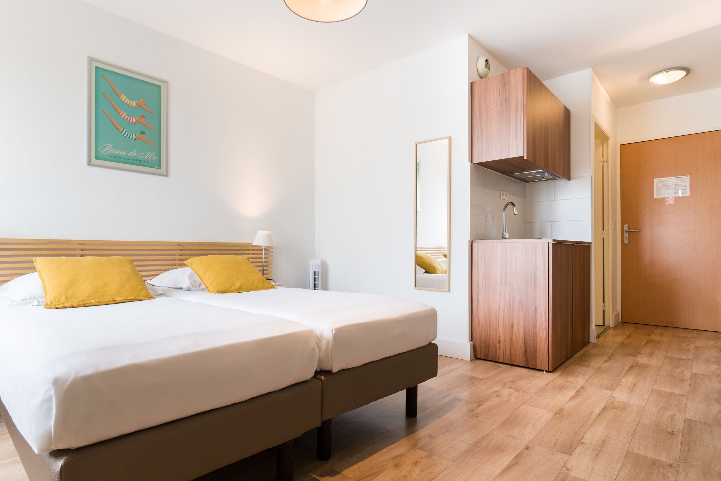 A hotel room with two beds, a wooden floor, a kitchenette, and a door.