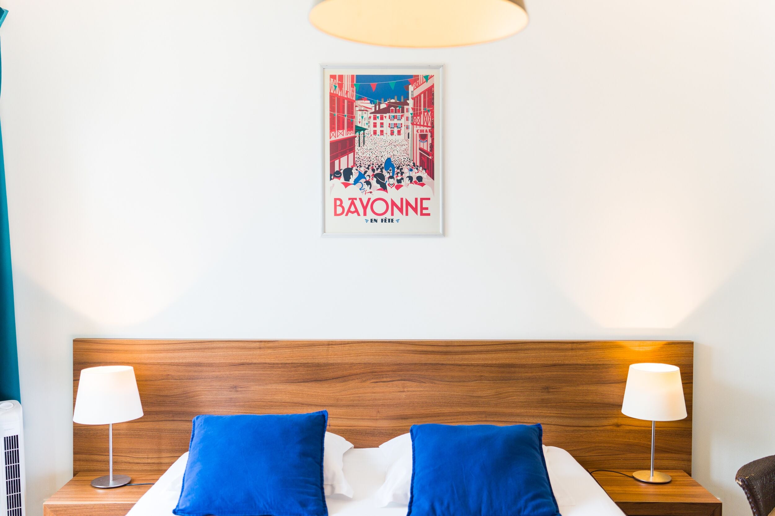 Habitación de hotel con cabecera de madera, dos almohadas azules, una colcha blanca y un cartel enmarcado en la pared.
