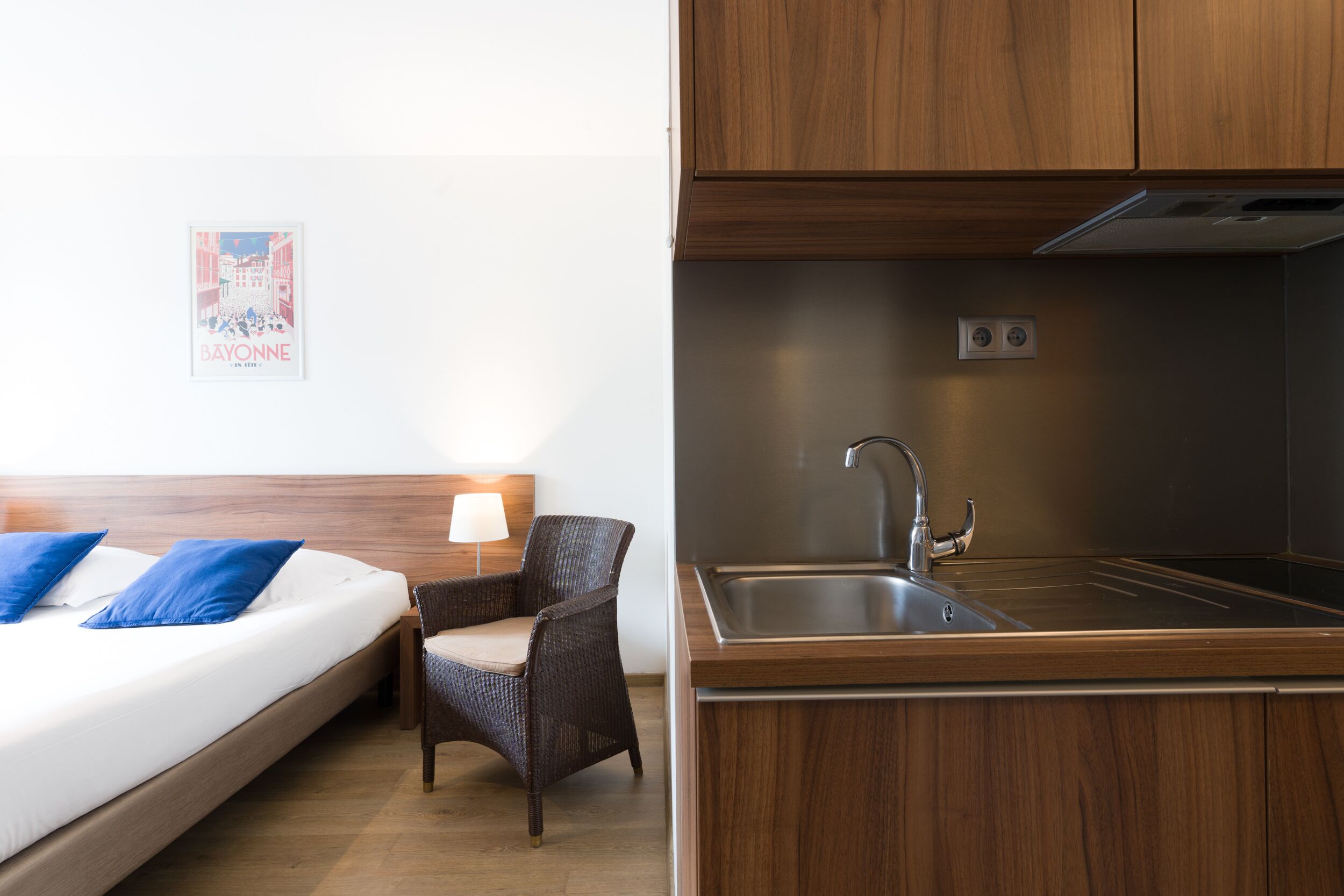 A hotel room with a bed, a chair, a nightstand, a wall-mounted lamp, and a poster.