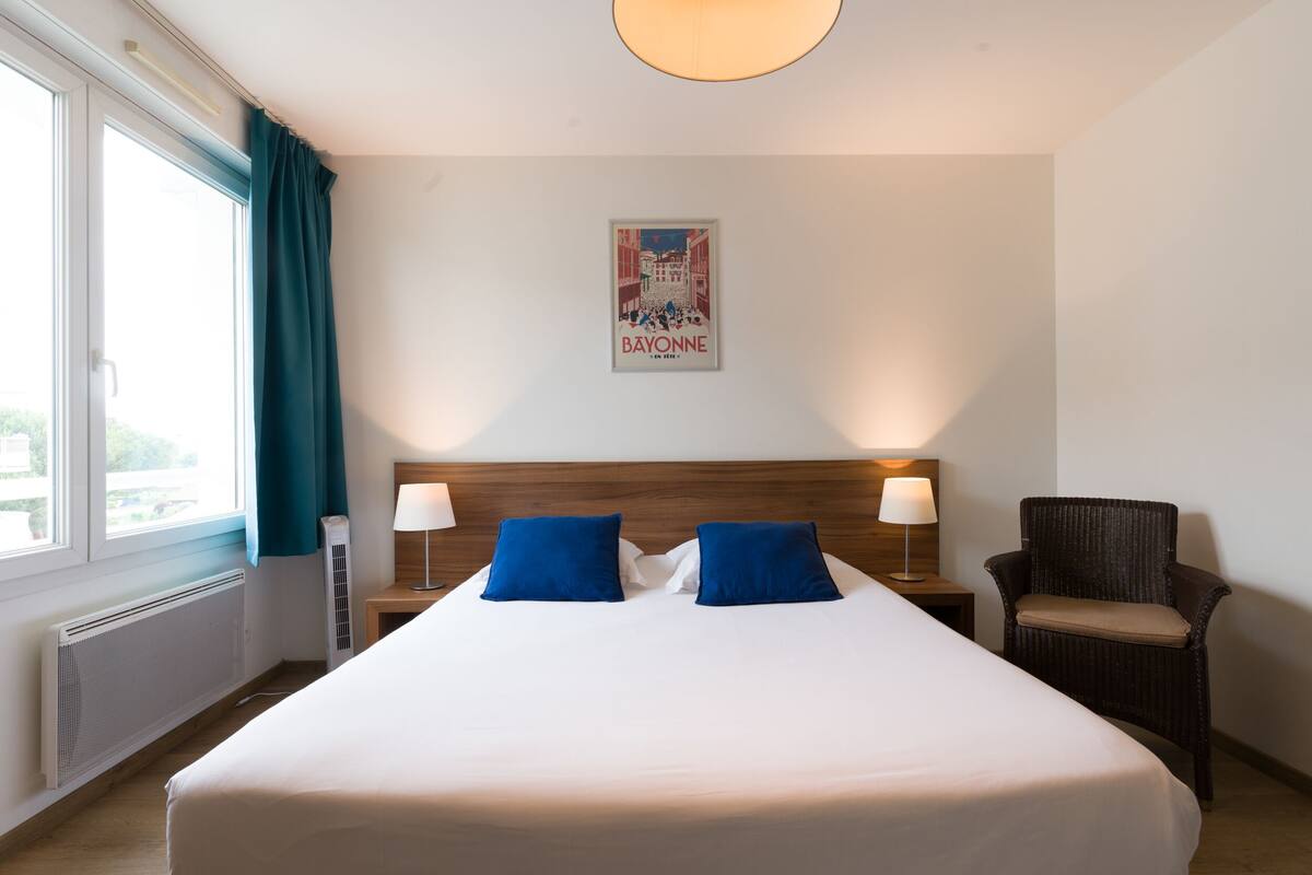 A hotel room with a large bed, two blue pillows, a wooden headboard, a wicker chair, a poster on the wall, and a window with curtains.