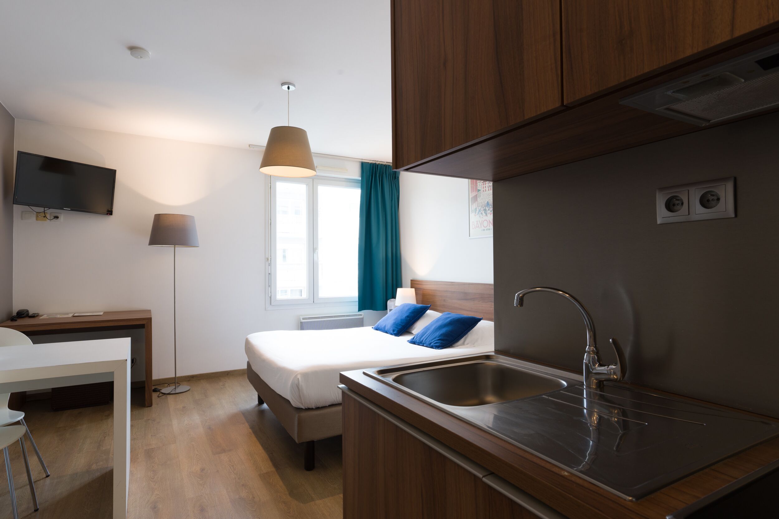A modern hotel room with a bed, a desk, a TV, a lamp, and a kitchen area with a sink.