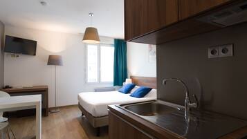 A modern hotel room with a bed, a desk, a TV, a lamp, and a kitchen area with a sink.