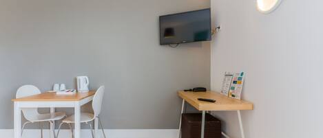 Una habitación pequeña con una mesa de madera, dos sillas blancas, un televisor montado en la pared y un escritorio con un teléfono y un revistero.