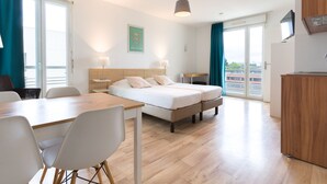 A modern hotel room with a wooden floor, a bed with two pillows, a desk with a chair, a small table with a lamp, and a TV on a cabinet. - Appart'Hotel Victoria Garden Pau (Pau)