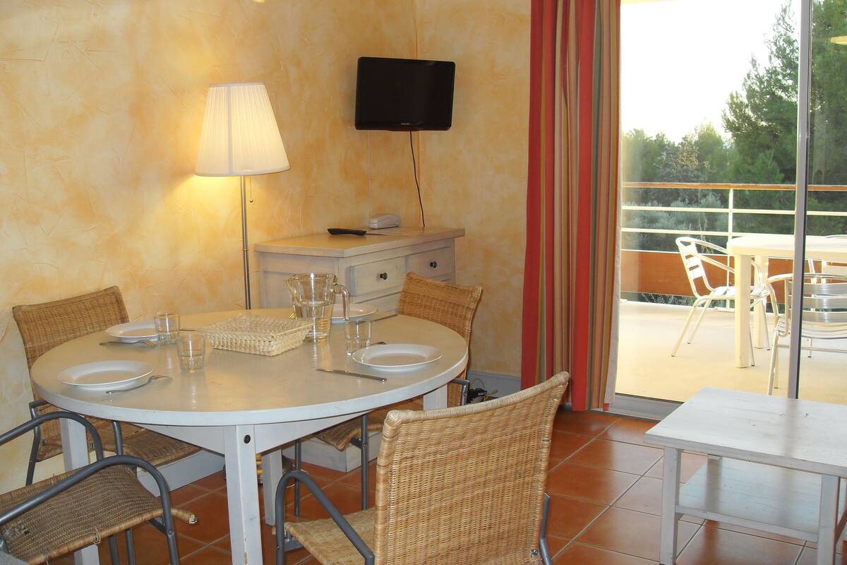 Un comedor con una mesa redonda, sillas de mimbre, una lámpara y un televisor en la pared.