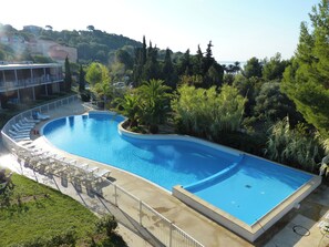 A large, curved swimming pool surrounded by lounge chairs and a deck area, with a view of a hillside and trees in the background. - Appart'hotel Victoria Garden La Ciotat (La Ciotat)