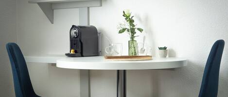 Um pequeno espaço de refeições com uma mesa branca, duas cadeiras azuis, uma máquina de café, um vaso com uma flor branca e uma prateleira com plantas e uma caixa.
