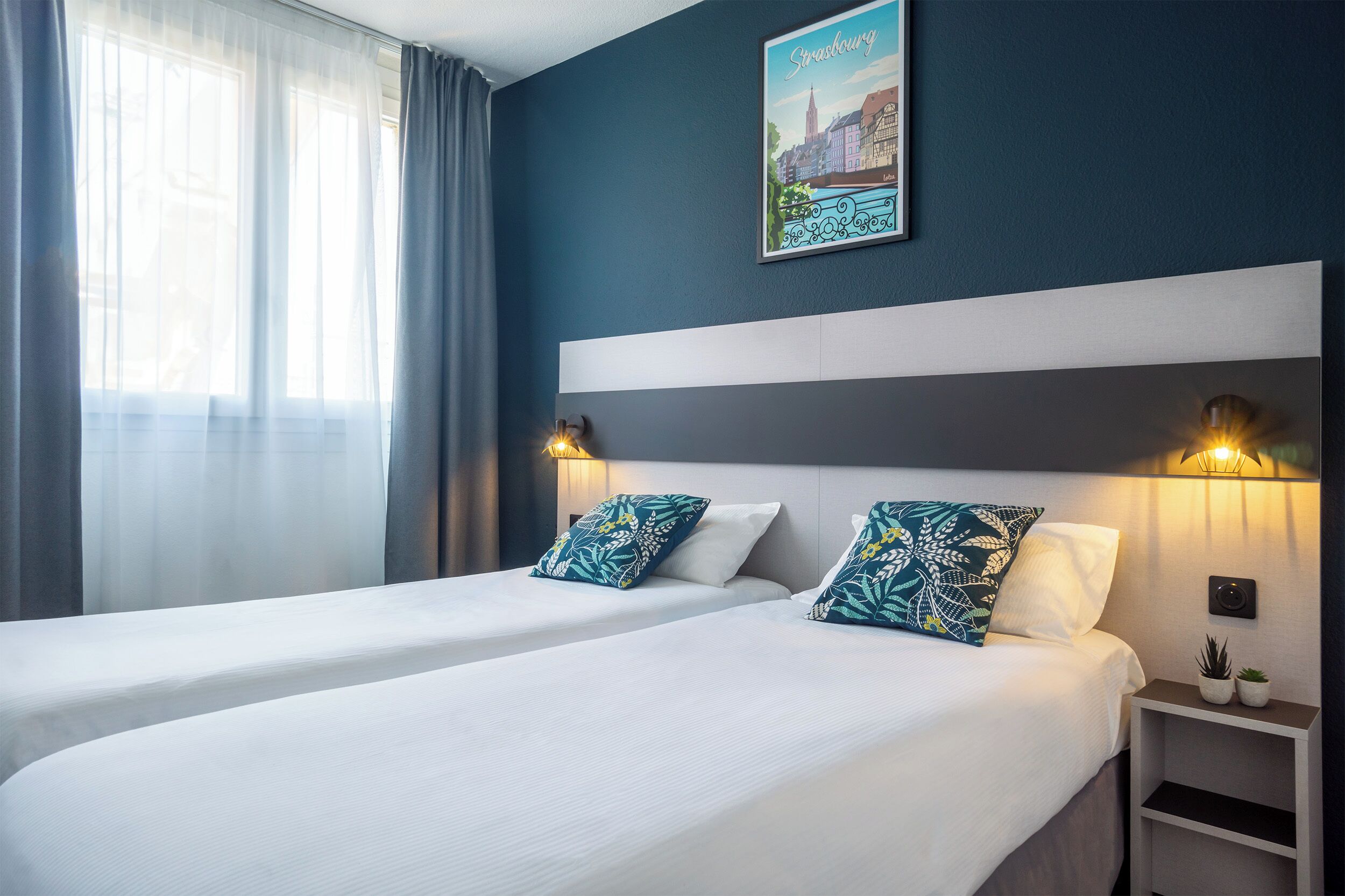 Una habitación de hotel con dos camas, una mesita de noche con lámpara, un cuadro con una vista urbana enmarcada y cortinas traslúcidas.