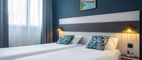 Una habitación de hotel con dos camas, una mesita de noche con lámpara, un cuadro con una vista urbana enmarcada y cortinas traslúcidas.