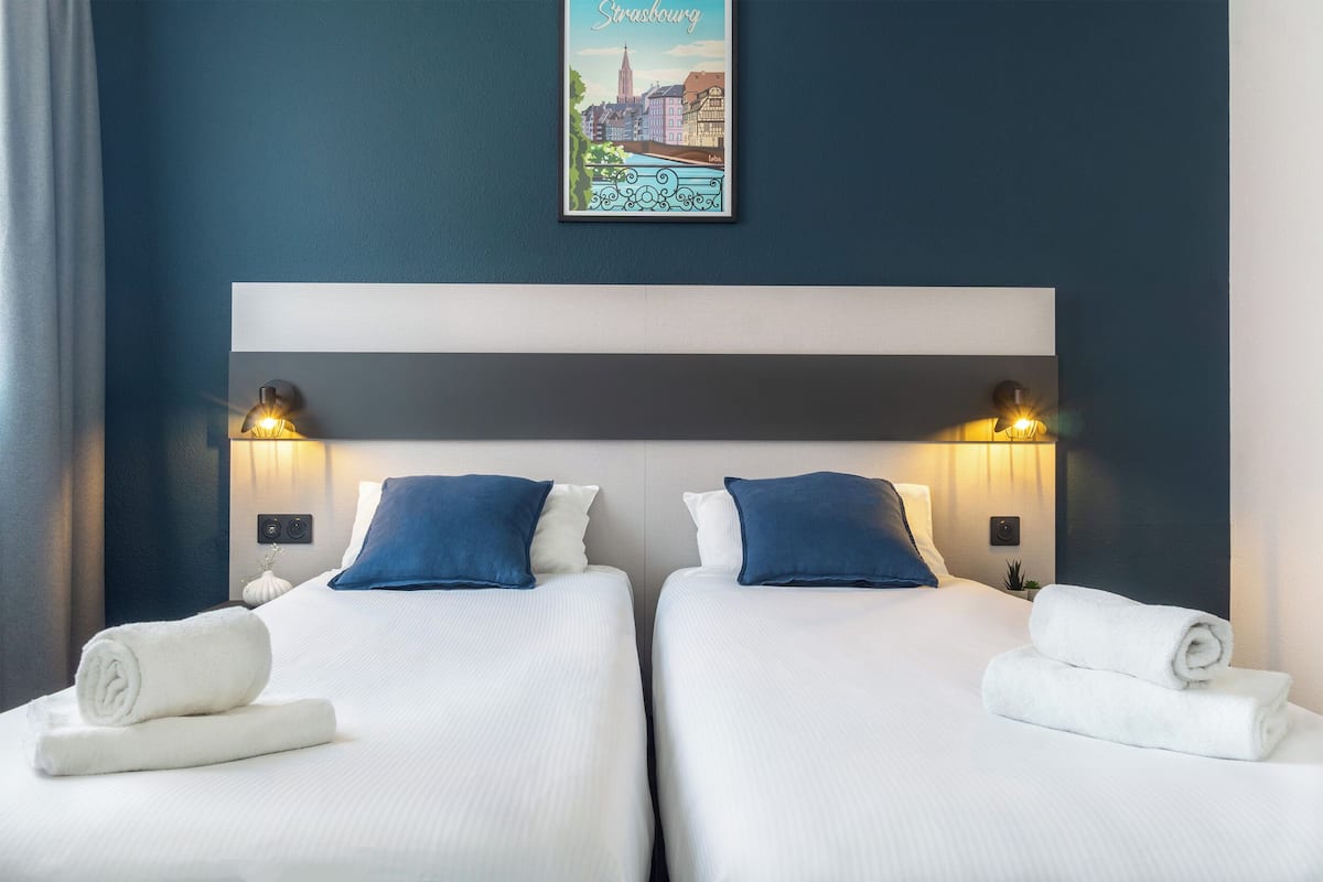 A hotel room with two beds, white linens, blue pillows, and a framed cityscape picture on the wall.