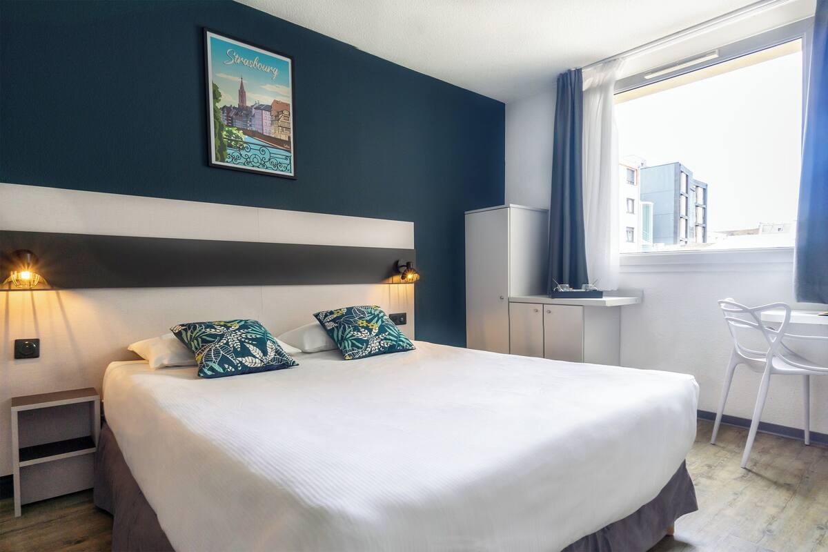 A hotel room with a bed, two bedside lamps, a nightstand, a desk with a chair, a window with curtains, and a cityscape poster on the wall.