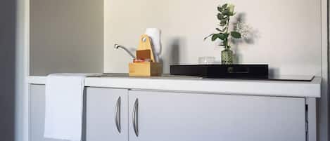 Une cuisine avec un plan de travail blanc, une planche à découper en bois, un couteau, un verre et un vase vert contenant une plante.