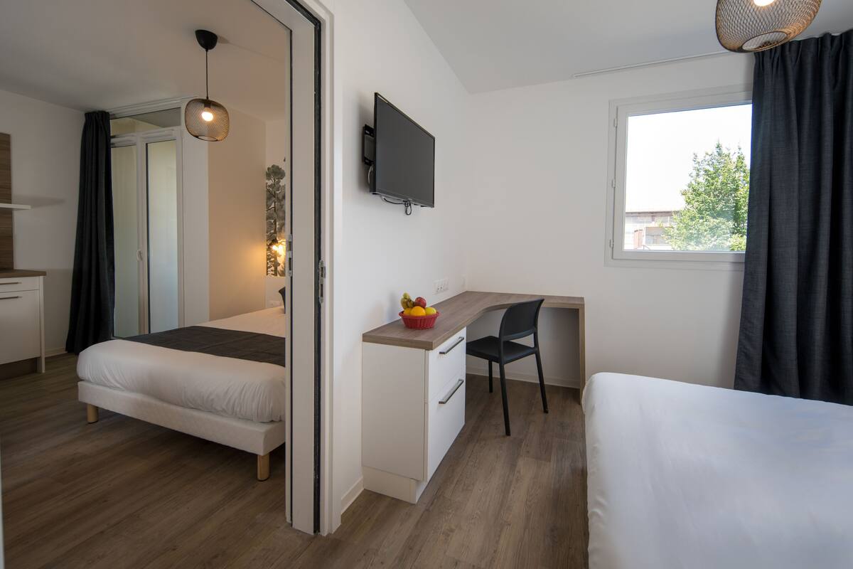 A bedroom with a bed, a desk with a chair, a television, and a window with curtains.