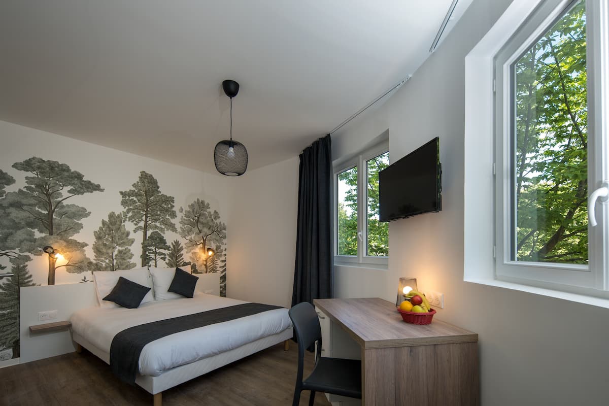 A bedroom with a bed, desk, chair, and a window with a view of trees.