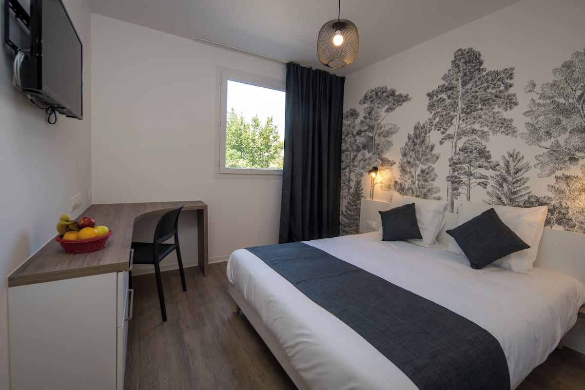 A bedroom with a bed, a desk with a basket of fruit, a chair, a window, and a wall with a tree mural.