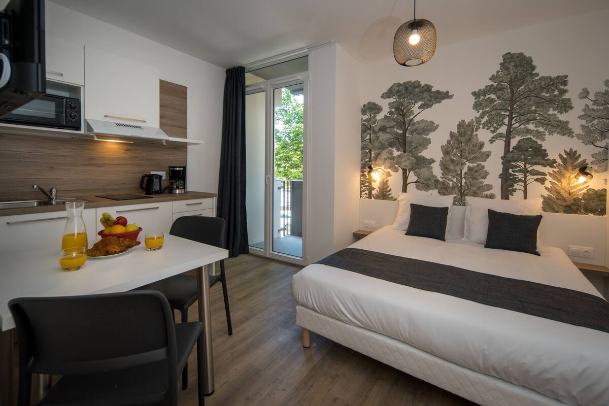 A modern room with a bed, a dining table with chairs, a kitchen area, and a large window with a view of trees.