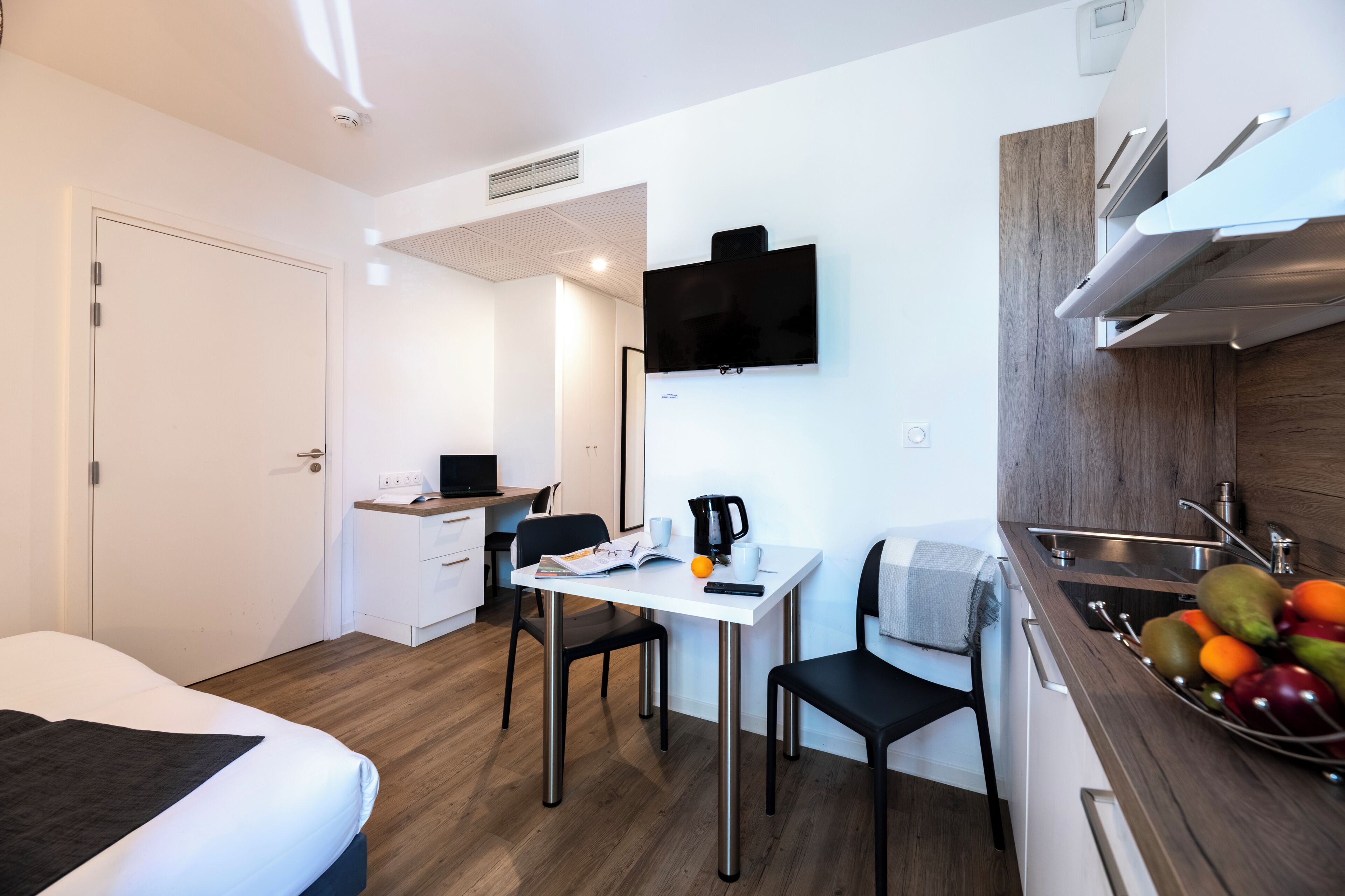 Una habitación de hotel compacta con cama, una kitchenette pequeña, un comedor con mesa y sillas, y un televisor montado en la pared.