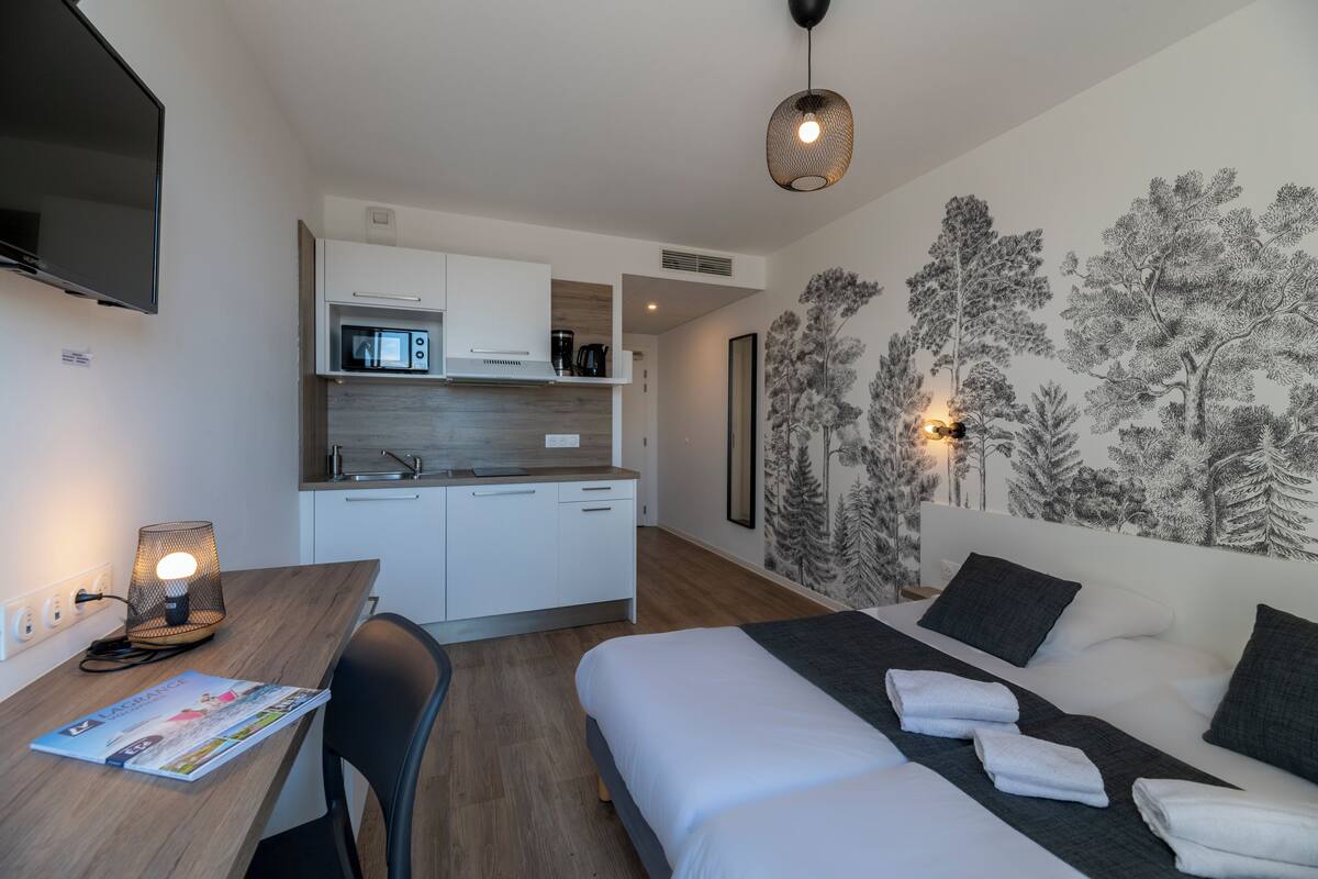A modern hotel room with a kitchenette, a bed with towels, a desk with a lamp, and a wall with a tree mural.
