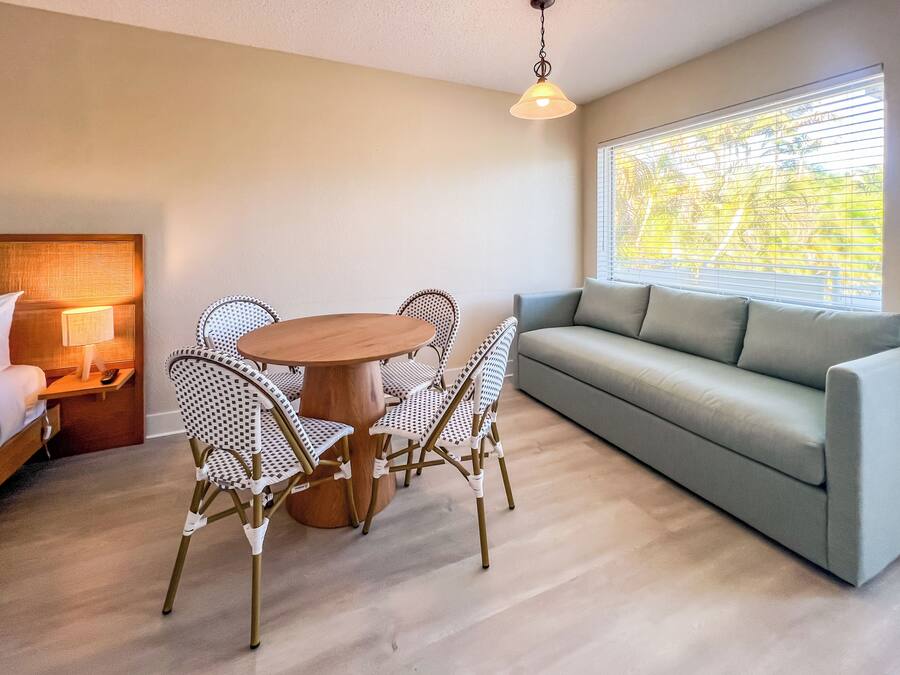 A modern living room with a round wooden table, patterned chairs, a grey sofa, and a window with blinds.