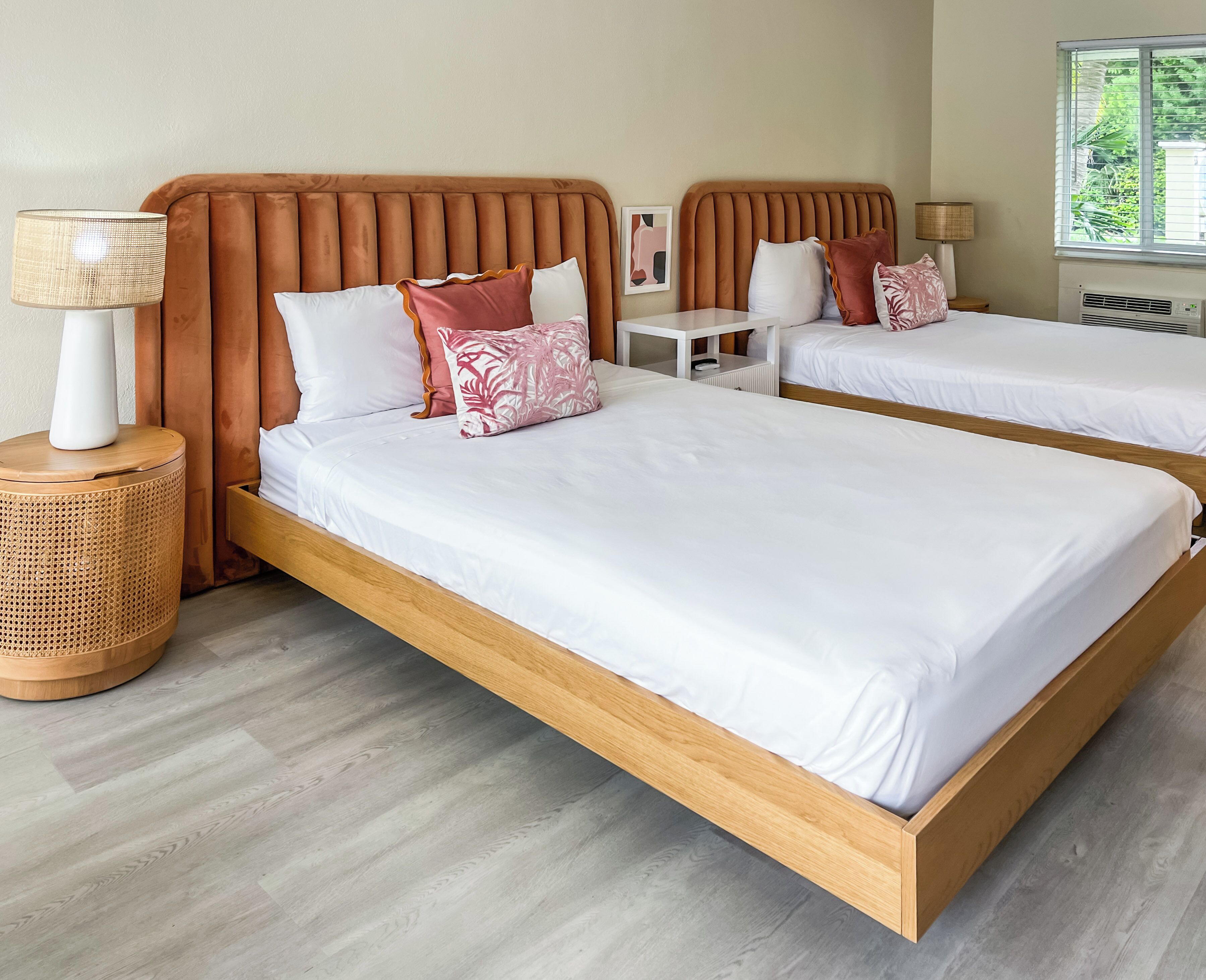 Habitación de hotel con dos camas, cada una con estructura de madera y ropa de cama blanca. Hay una mesita de noche con una lámpara y una cesta tejida en el suelo, junto a la cama.