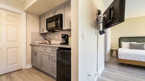 Une cuisine compacte équipée d’un four à micro-ondes, d’une machine à café et d’un lave-vaisselle. À côté se trouve une chambre avec un lit et une table de chevet.