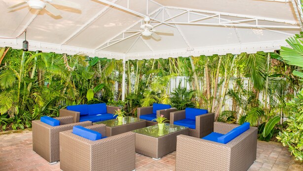 Un espace extérieur aménagé en terrasse, avec des meubles en osier et une table à plateau de verre, le tout entouré d’une végétation tropicale luxuriante.
