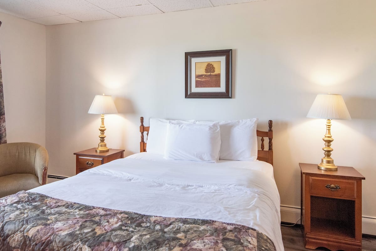 A hotel room with a bed, two bedside tables with lamps, a chair, a framed picture on the wall, and a window with curtains.
