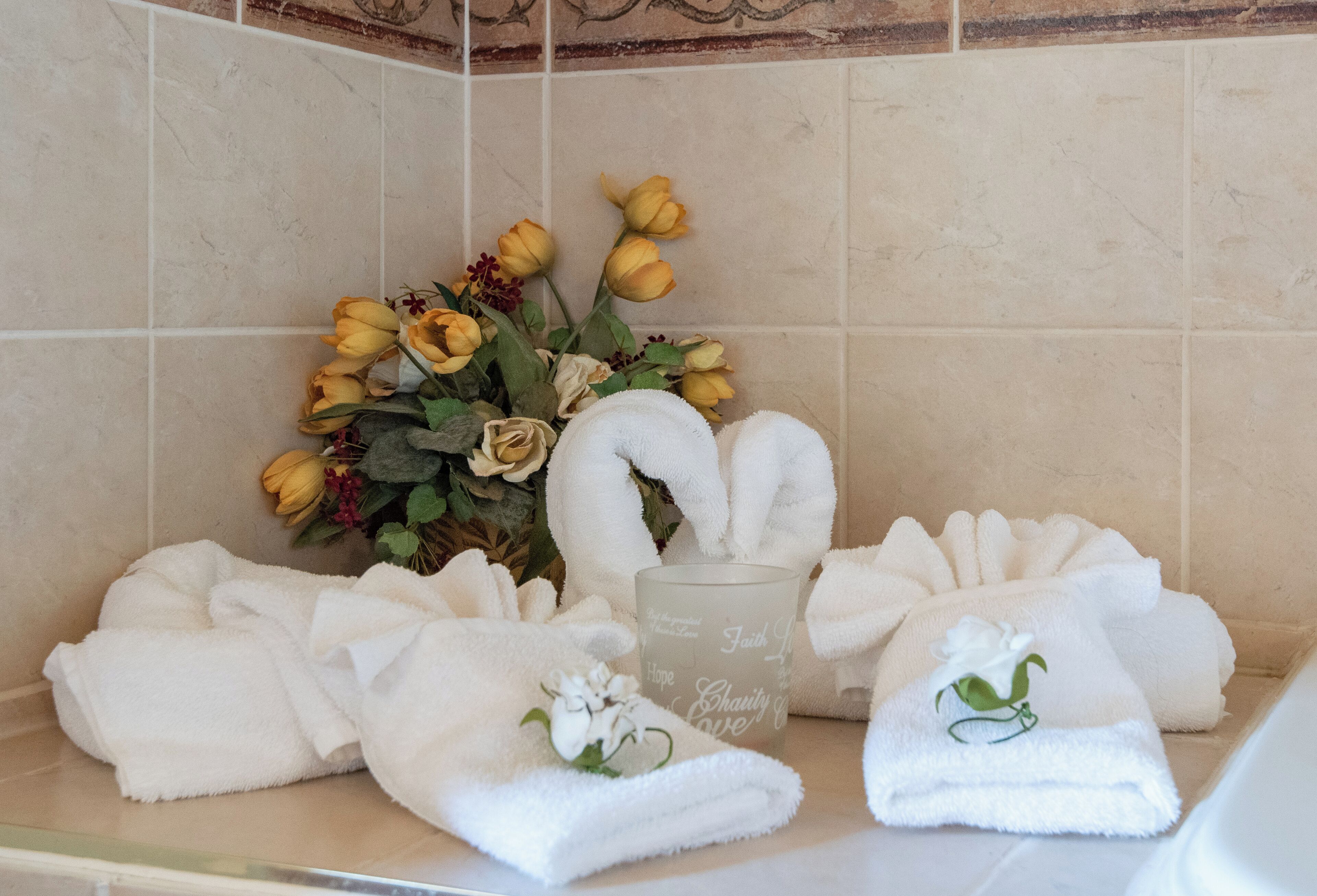 A bathroom with a towel rack holding several white towels, a glass candle with inscriptions, and a bouquet of yellow and white flowers.
