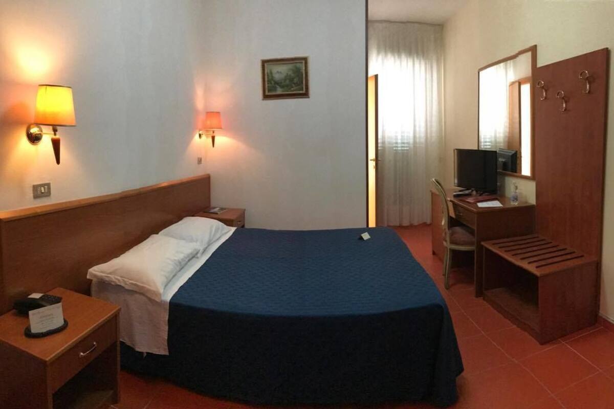Una habitación de hotel con cama, mesita de noche, lámpara, escritorio, silla y armario.