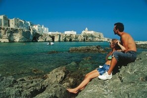Una pareja sentada sobre rocas junto al mar, con edificios al fondo.