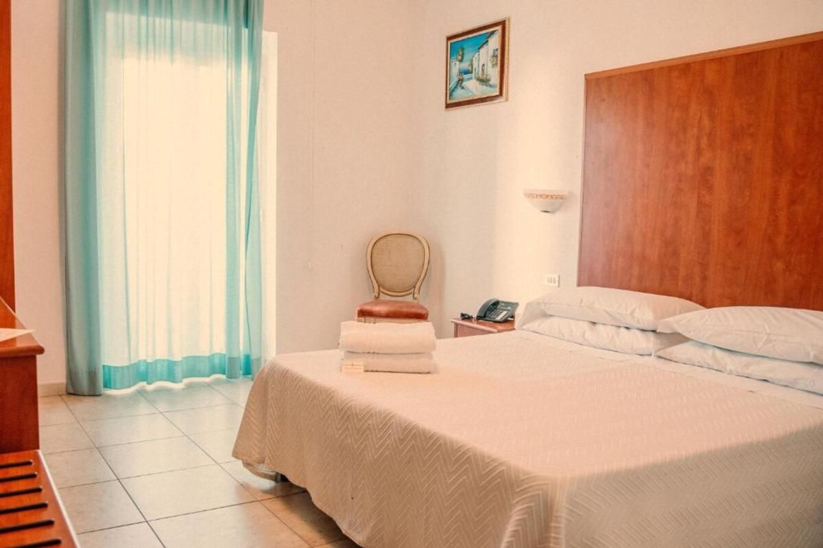 A hotel room with a bed, a chair, a small table, a telephone, and a painting on the wall.