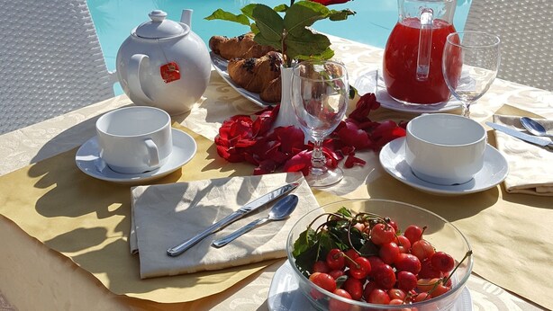 Une table placée au bord de la piscine, avec une théière, des tasses, un verre contenant une boisson rouge, une assiette de croissants, un vase avec une rose rouge et un bol rempli de baies.