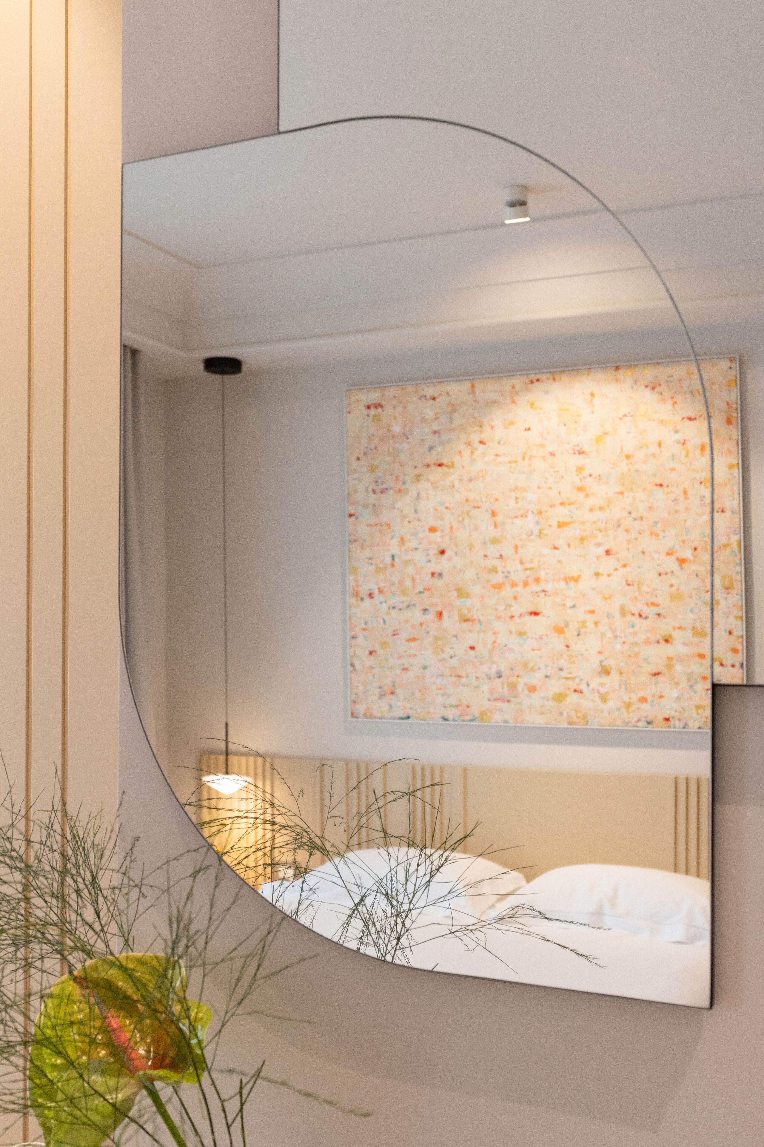 A modern hotel room with a large mirror reflecting a colorful abstract painting on the wall, a neatly made bed, and a plant in the foreground.