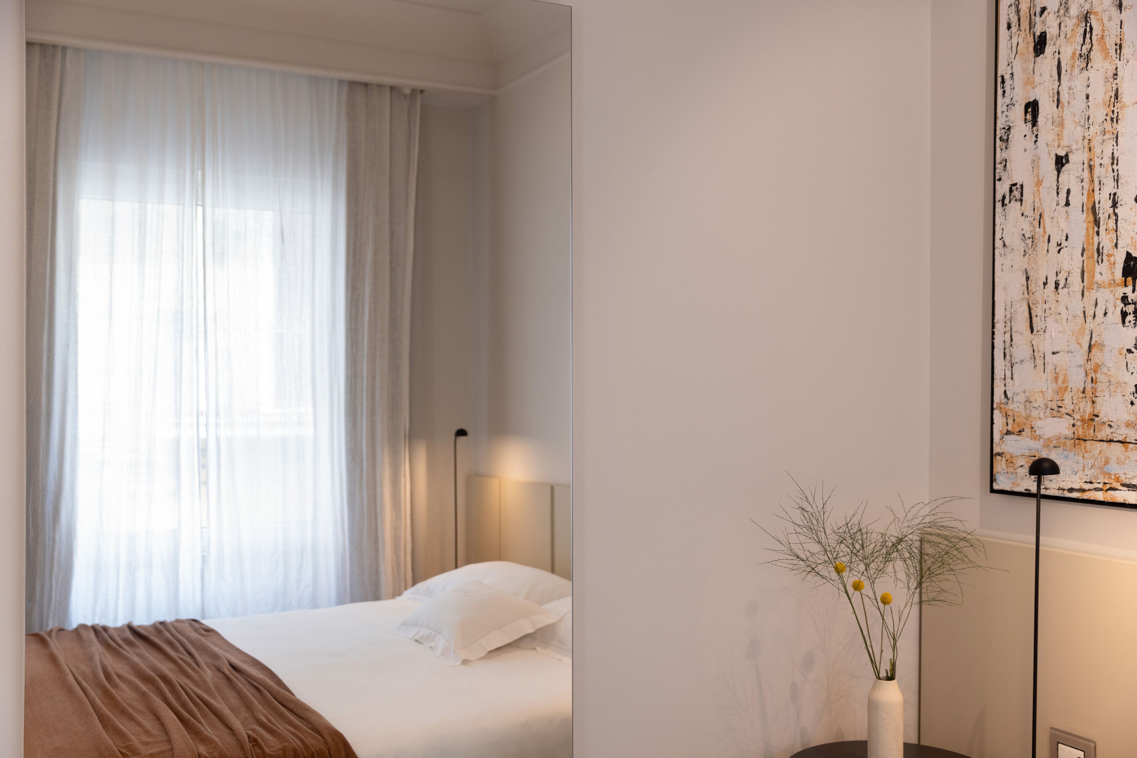 A hotel room with a bed, a bedside table, a lamp, a window with sheer curtains, and a wall-mounted artwork.