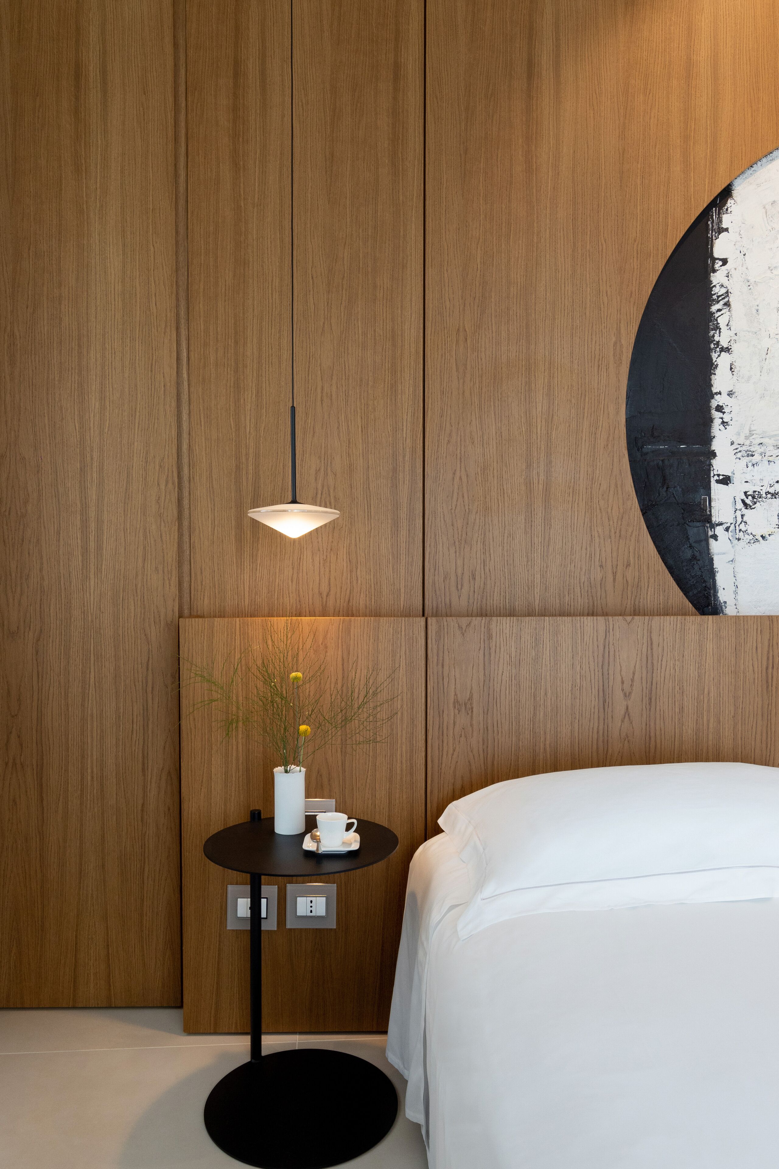 A modern hotel room with a wooden panel wall, a round black table with a white vase and a cup, and a bed with white bedding.