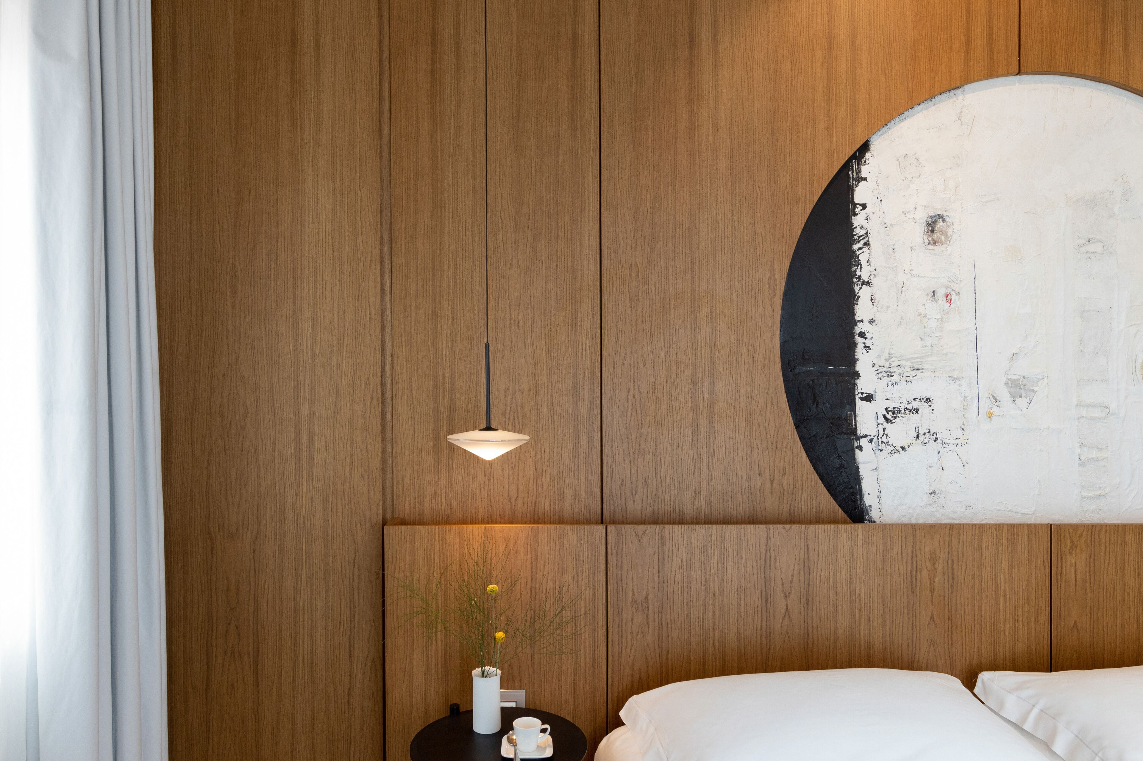 A hotel room with a wooden panel wall, a round artwork, a white bed, and a small table with a vase and a cup.