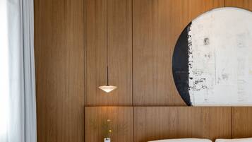 A hotel room with a wooden panel wall, a round artwork, a white bed, and a small table with a vase and a cup.