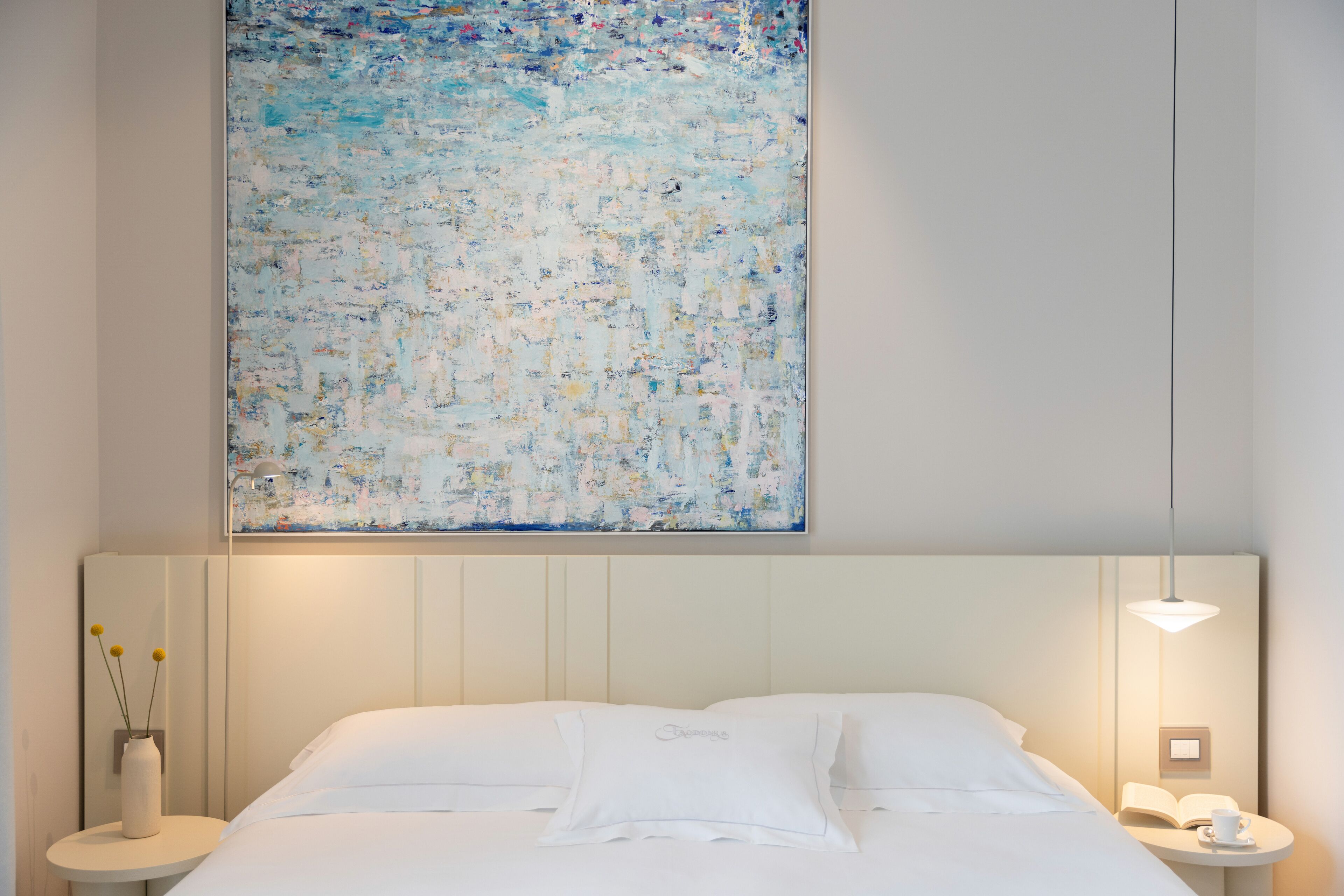 Ein Hotelzimmer mit einem großen abstrakten Gemälde, einem ordentlich bezogenen Bett, einem Nachttisch mit Lampe und einem kleinen Tisch mit Vase und Buch.