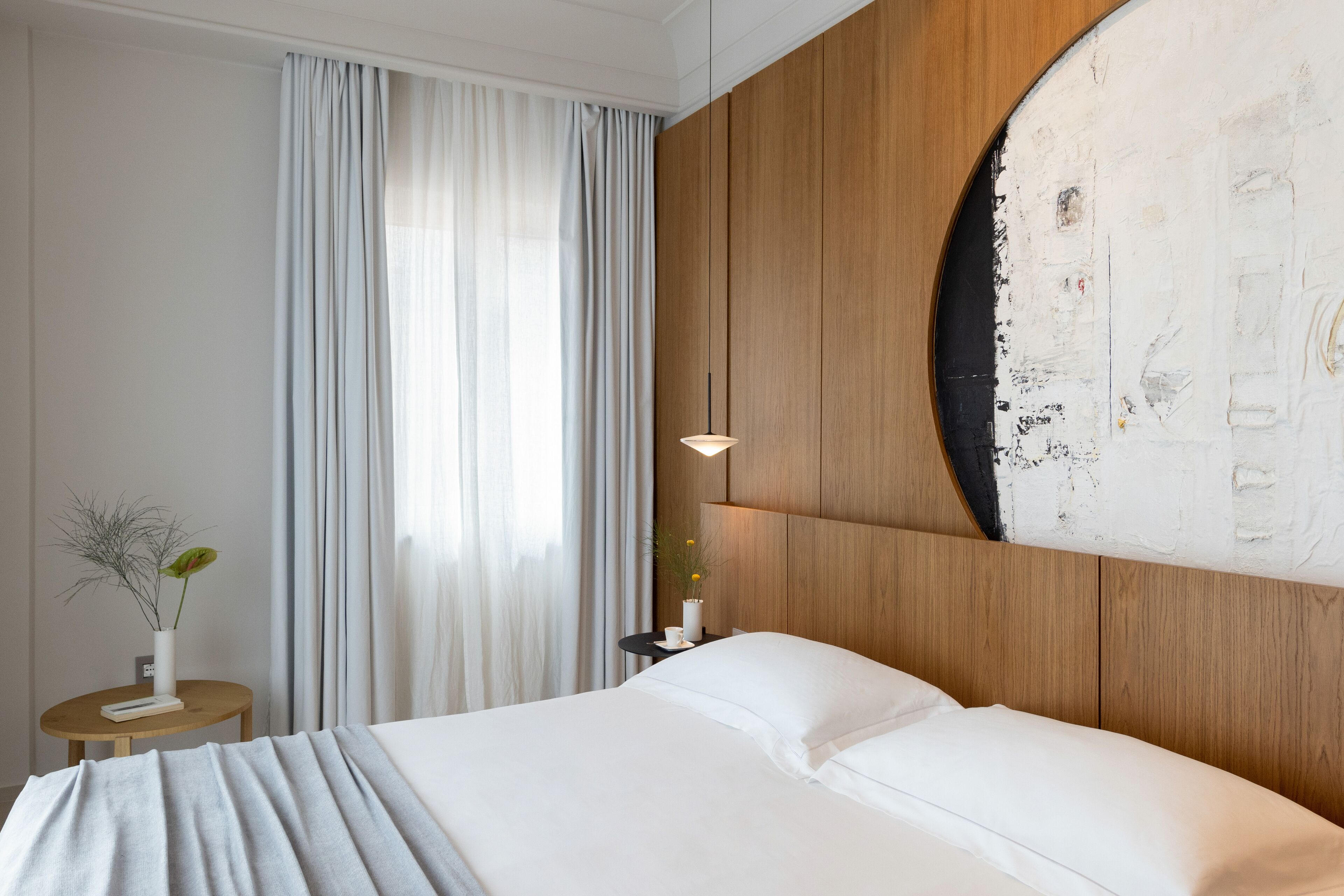 A hotel room with a large bed, a wooden headboard, a round painting, a nightstand with a lamp, and a window with curtains.