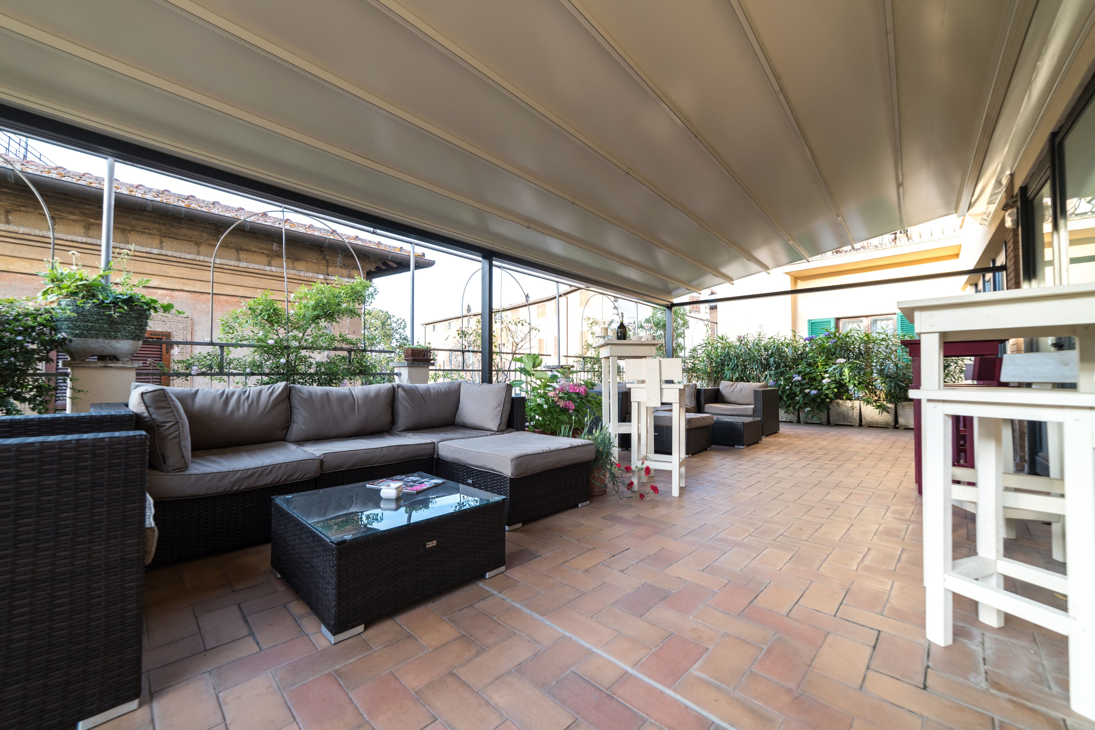 Een terras met een loungeset, een salontafel met glazen blad, rieten meubels en potplanten.