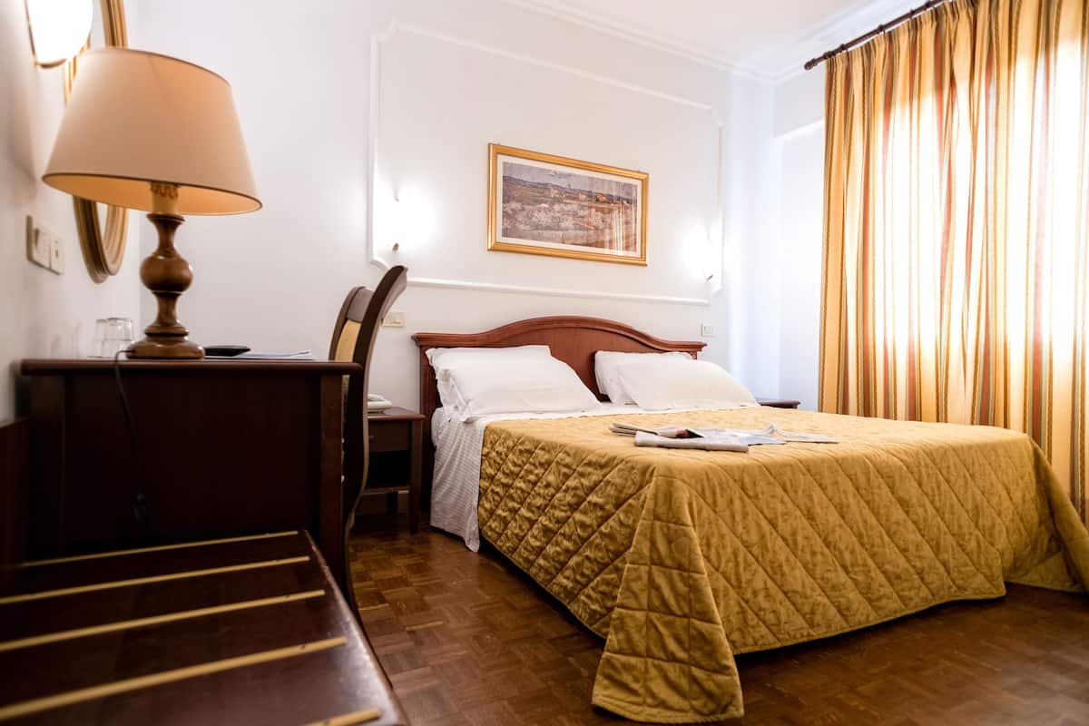 Une chambre d’hôtel avec un lit, un bureau avec une lampe, une chaise, un tableau encadré au mur et une fenêtre avec des rideaux.